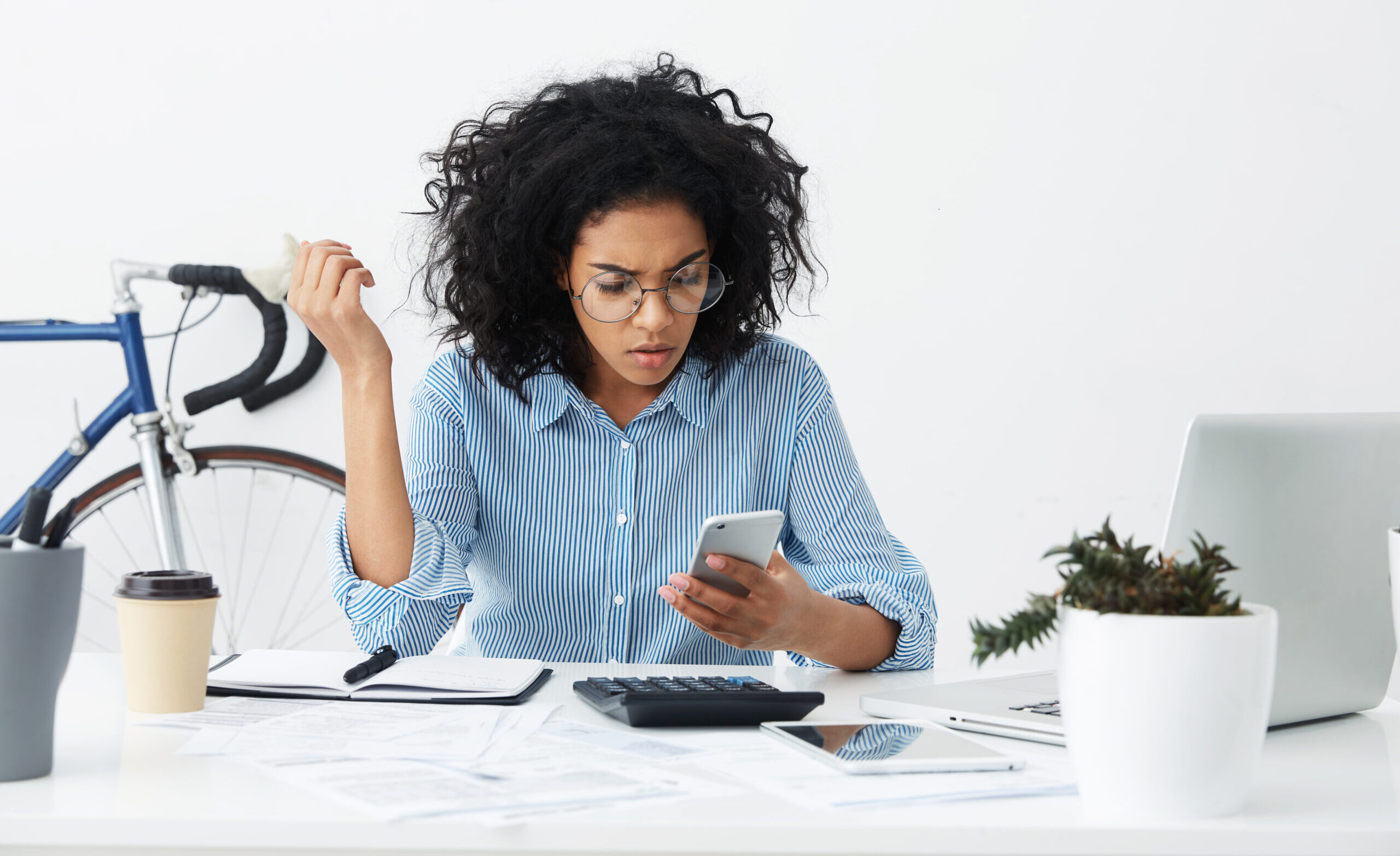Perplexed young woman employee in formal shirt and round eyewear having frustrated look while facing a problem with mobile phone during working day in office, doing paperwork. People and technology
